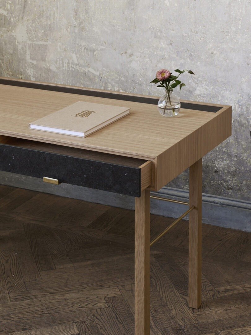 A wooden desk with a black drawer, a closed book, and a small vase with a pink flower on top. The background is a textured wall, and the floor is dark wood.