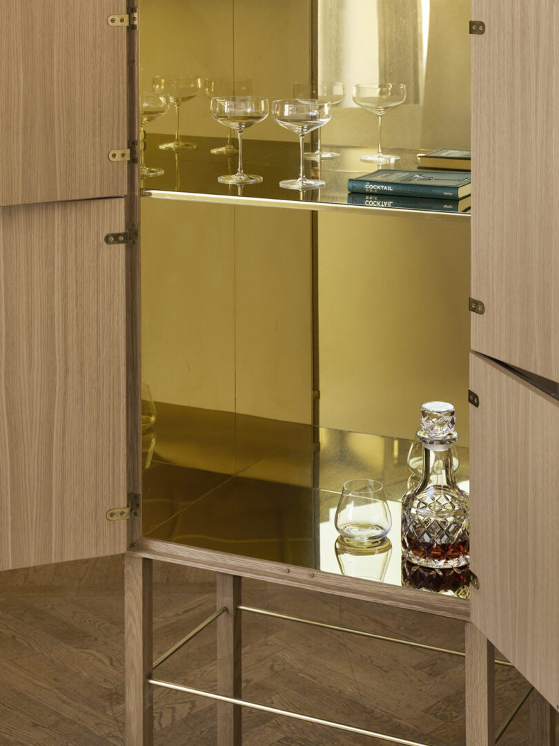 Open wooden cabinet with glass shelves, holding several empty glass goblets, a few books, a decanter, and a tumbler with liquid inside.