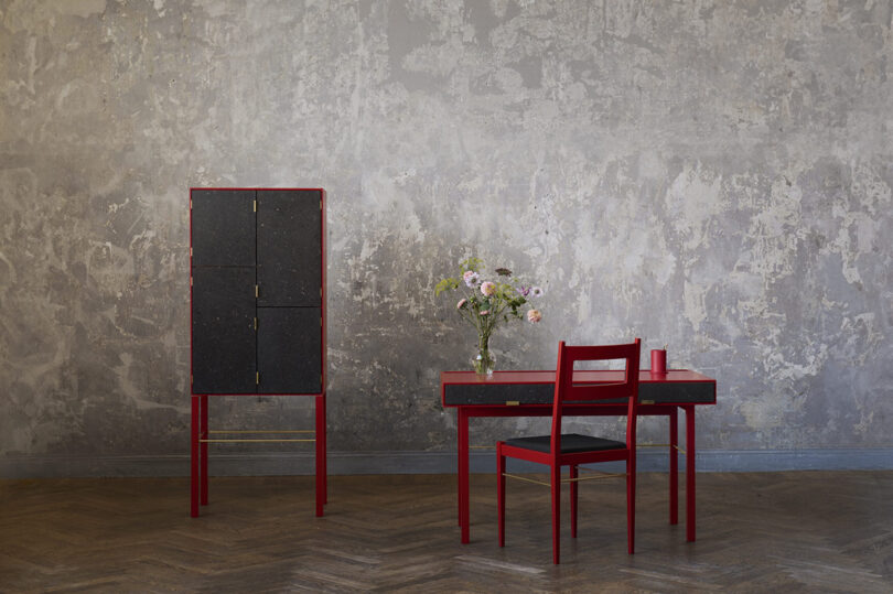 A red and black furniture set with a cabinet, table, and chair stands against a textured grey wall, with a vase of flowers on the table.