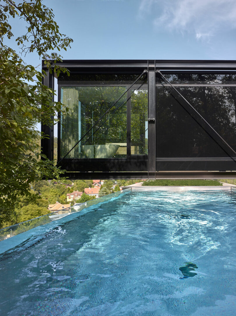 A modern, glass-walled building extends over a pool with clear blue water, surrounded by trees and overlooking a scenic view.