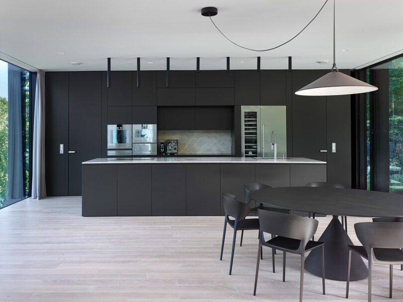 Modern kitchen with black cabinetry, a sleek island with built-in appliances, and a round dining table with matching chairs. Large windows offer a view of greenery outside.