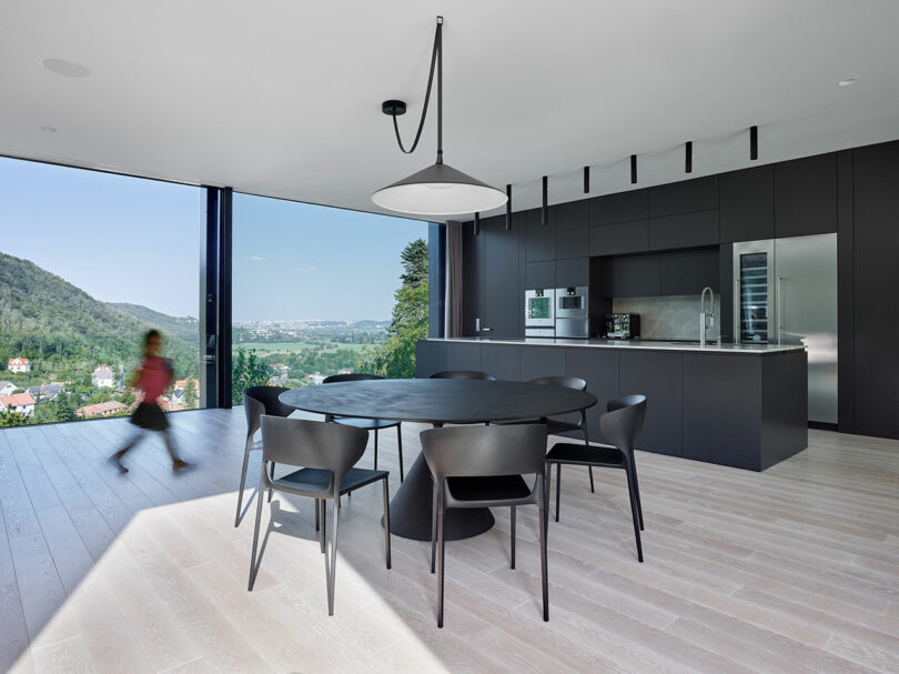 Modern kitchen with black cabinetry and a large round table. Floor-to-ceiling windows showcase a landscape view. A child runs by the window.