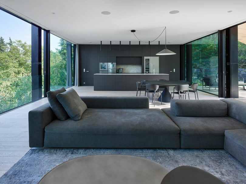 Spacious modern living room with a large gray sectional sofa. Open kitchen area in the background, featuring dark cabinetry and a dining table. Floor-to-ceiling windows offer a view of greenery.