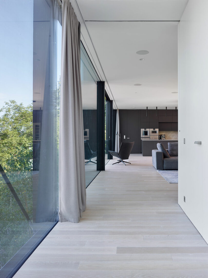 Modern interior with large glass windows, light wood flooring, gray curtains, and a view of trees. A gray sofa and armchair are near a kitchen area with dark cabinets.