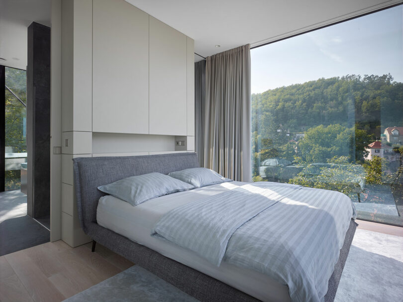 Modern bedroom with a large bed, light bedding, and a floor-to-ceiling window offering a view of trees and houses outside.