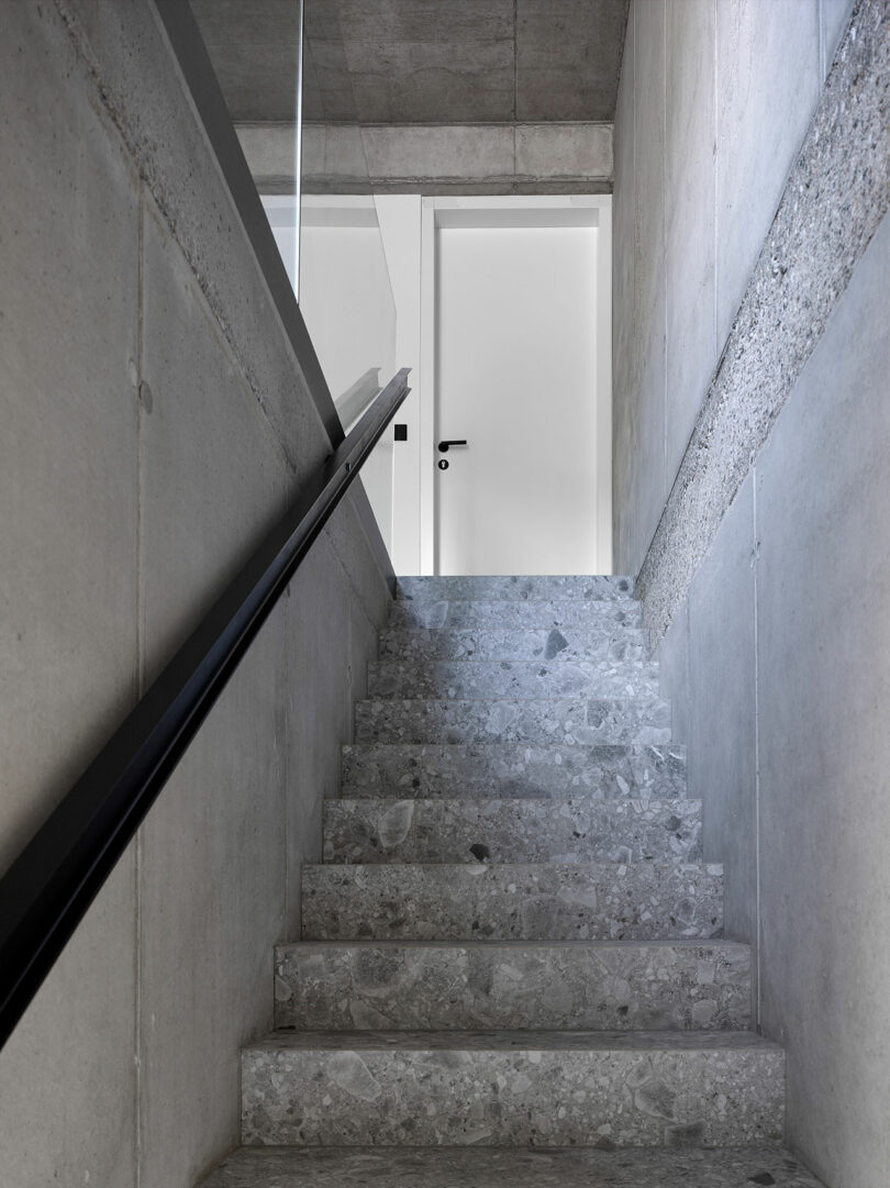 Concrete staircase with a black handrail leading to a closed white door, surrounded by gray walls with a textured middle section.