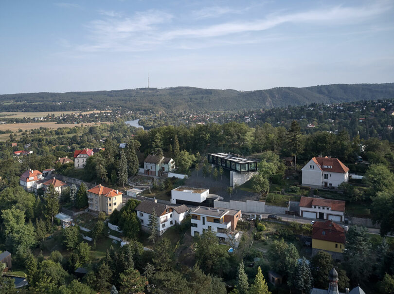 Aerial view of a suburban area with scattered houses, surrounded by trees. A river and hills are visible in the background.