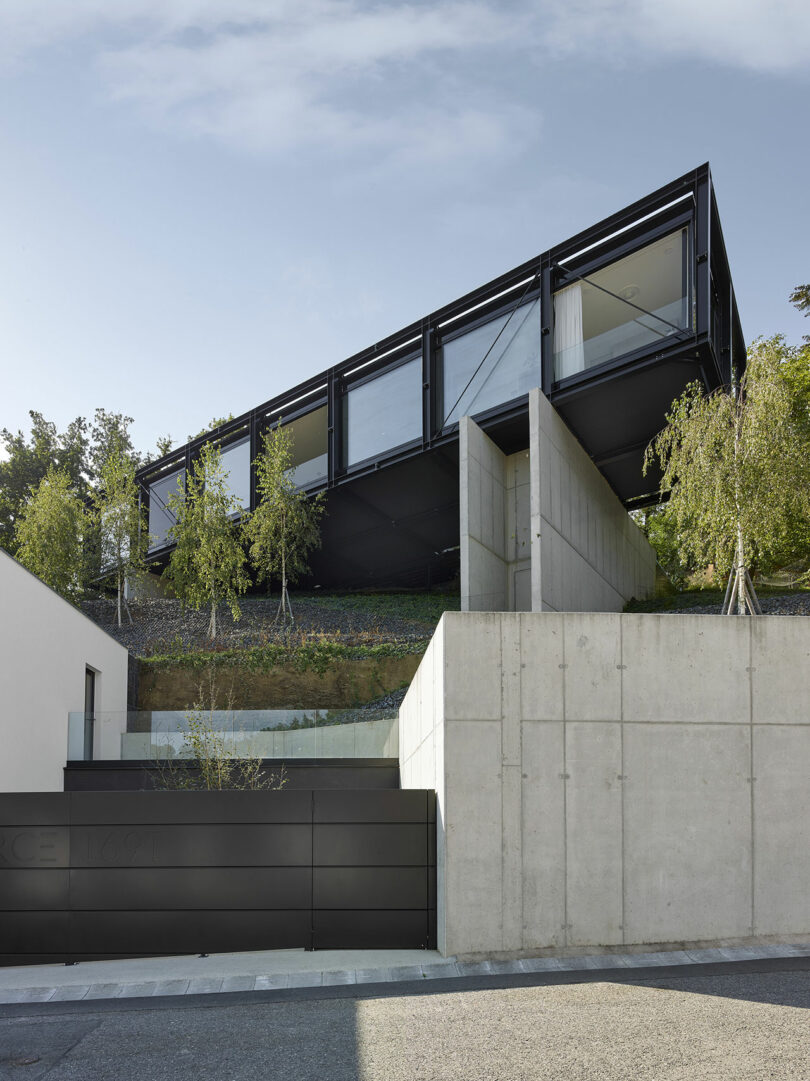 Modern house with a sleek black geometric design, elevated on a hillside above a concrete wall. Trees are planted around the structure.