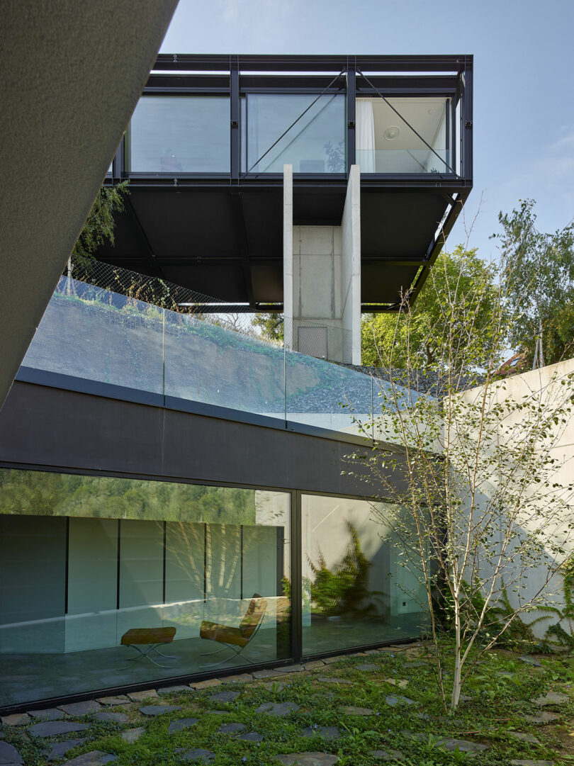 Modern house exterior with large glass windows, elevated section supported by columns, and surrounding greenery.