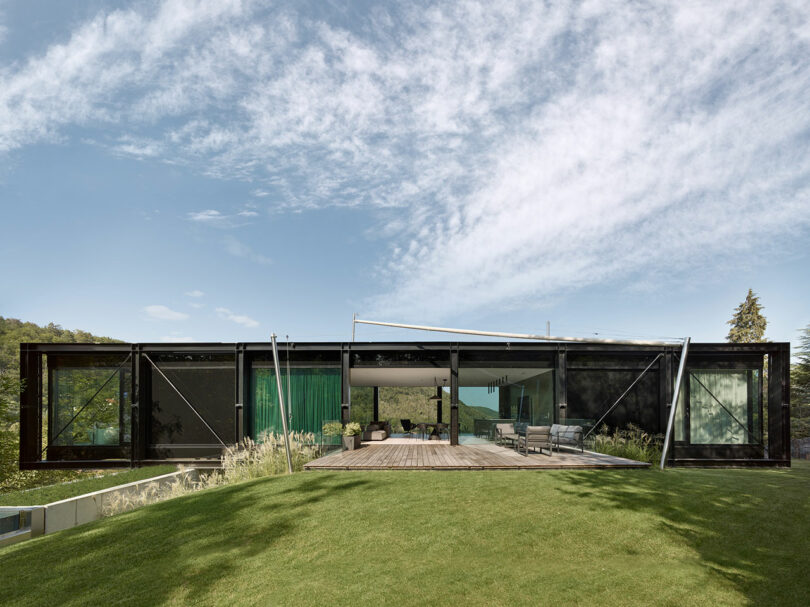 Modern glass and steel house with a large wooden deck set on a grassy lawn, surrounded by trees under a partly cloudy sky.