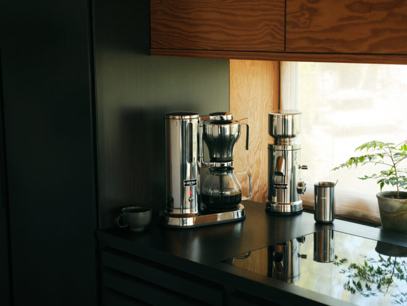 The Aarke Coffee System sits elegantly on a black kitchen counter, accompanied by a sleek grinder. Nearby, a small plant adds a touch of greenery beside the inviting mug, creating the perfect coffee nook.