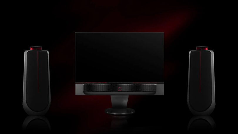 A sleek computer monitor with two tall, black speakers on each side, set against a dark background with a red accent.