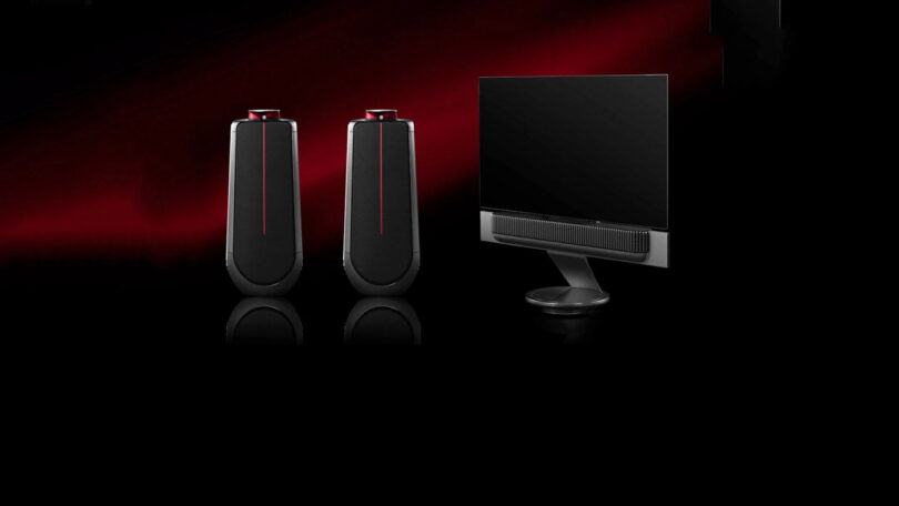 A pair of sleek black speakers with a red accent are placed next to a modern flat-screen computer monitor on a black and red gradient background.