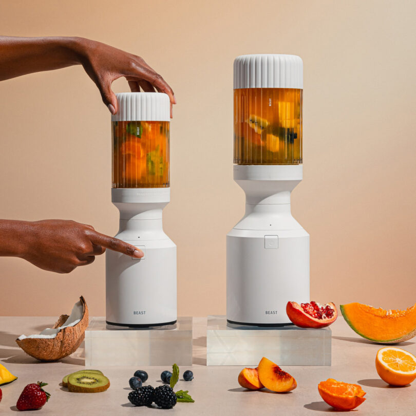 Two hands operate white blenders filled with fruit on a tabletop. Surrounding the blenders are cut fruits like oranges, berries, and a coconut.