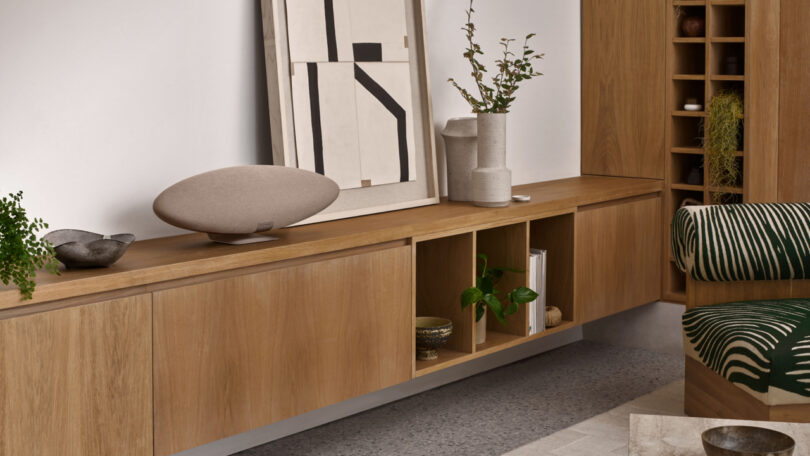 Modern living room with a wooden sideboard, abstract artwork, plants, and a beige Zeppelin Pro Edition speaker. A green patterned chair is partially visible in the foreground, adding a touch of style to the contemporary space.