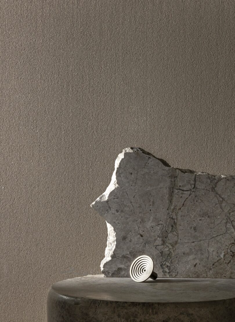 A stone slab stands upright on a smooth, dark surface with a small, spiral knob at its base, set against a textured neutral wall.