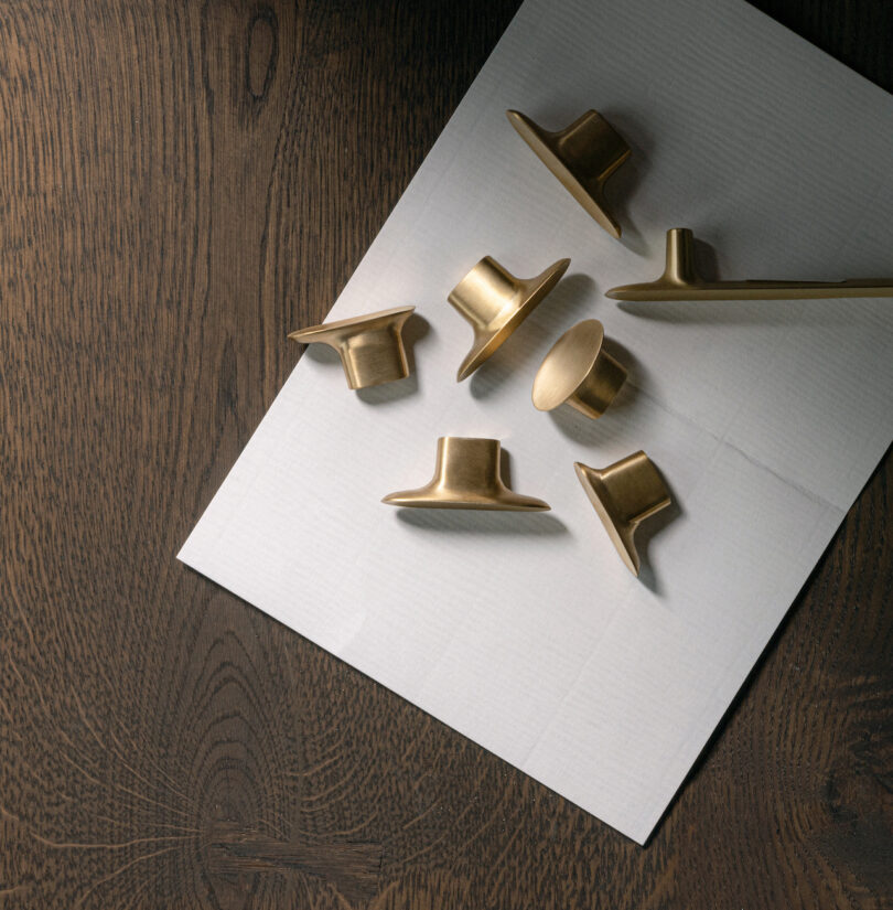 Assorted brass cabinet knobs shaped like hats and cylinders are arranged on a textured white paper, set against a dark wooden surface