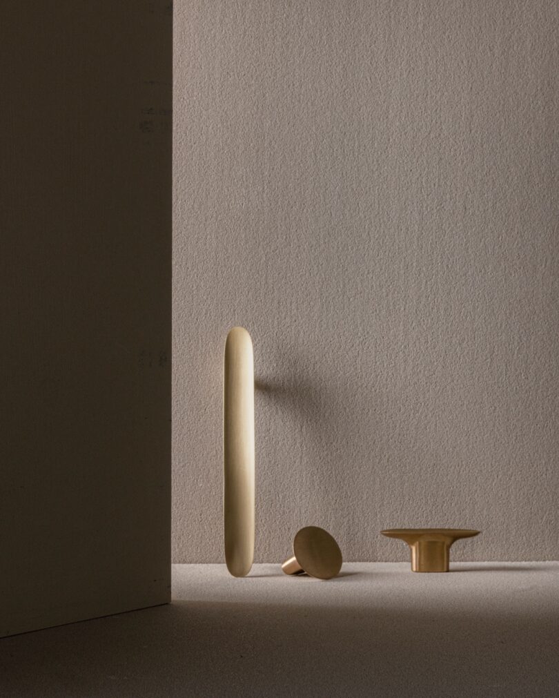 Three wooden objects arranged against a textured beige wall: a tall narrow oval, a round disc leaning at an angle, and a short, wide pedestal