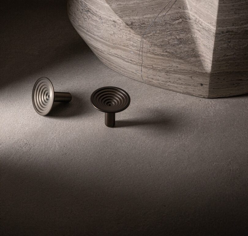 Two round, metallic, spiral-patterned knobs on a textured stone surface next to a large, geometric stone structure.