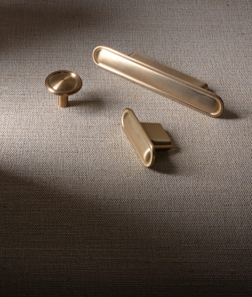 Three brass drawer pulls of varying shapes are displayed on a textured fabric background