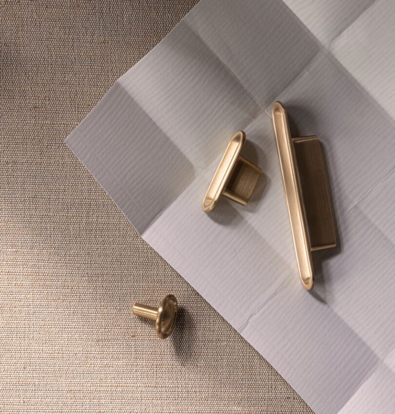 Three gold cabinet handles on a textured beige surface with a white folded paper backdrop