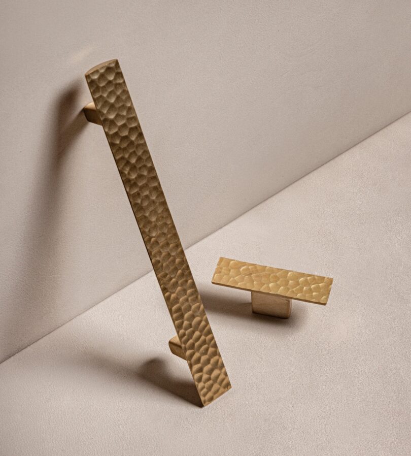 Two textured gold cabinet handles are positioned on a beige surface, one vertical and one horizontal