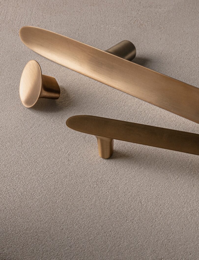 Three bronze-colored, smoothly textured cabinet handles of different shapes and sizes on a beige textured surface