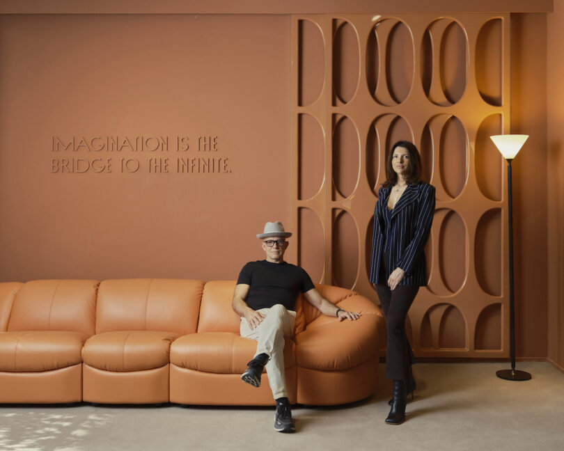 A man and woman are posing next to an orange sofa in a room with an orange backdrop and a lamp. The wall features the text "Imagination is the bridge to the infinite"