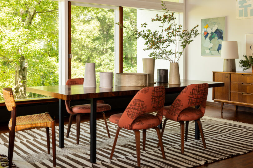 A dining room with a wooden table, six orange cushioned chairs, a zebra-striped rug, and plants. Large window offers a view of greenery outside