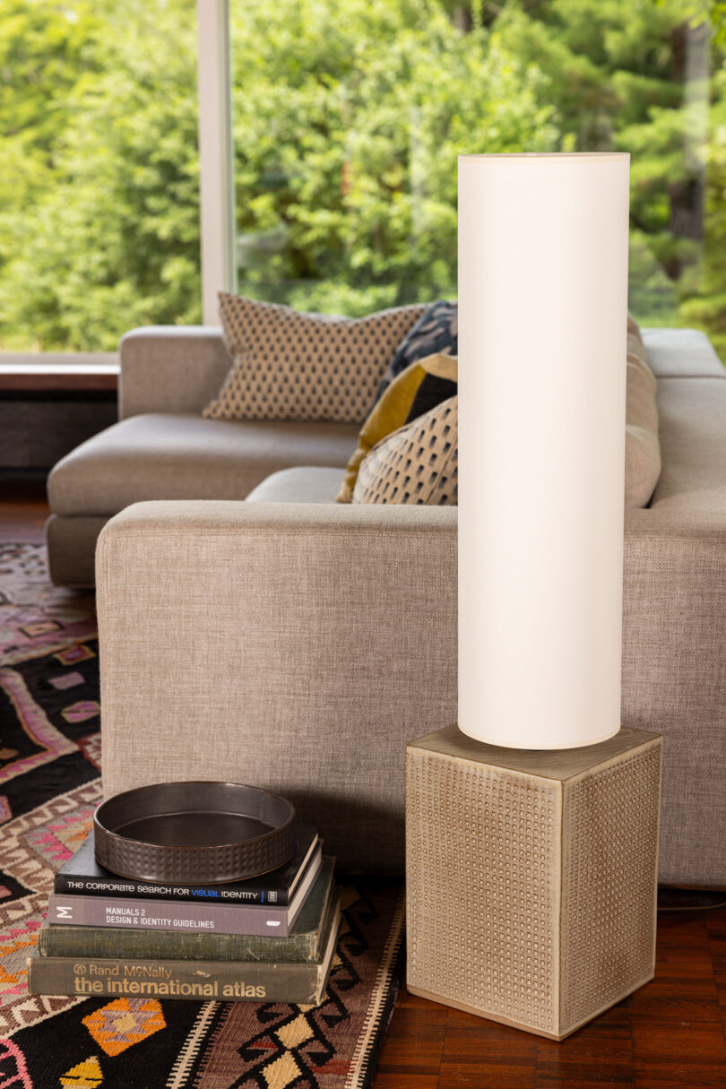 A modern living room featuring a gray couch, patterned cushions, a white cylindrical lamp on a textured base, a stack of books including "The International Atlas," and a round metal tray on a rug