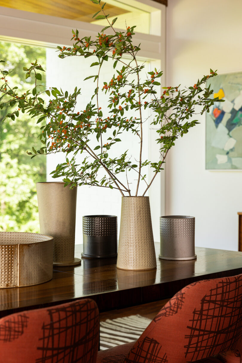 A modern dining table with red-cushioned chairs features a tall vase with branches and small orange berries, surrounded by other decorative containers. A window and abstract artwork are in the background