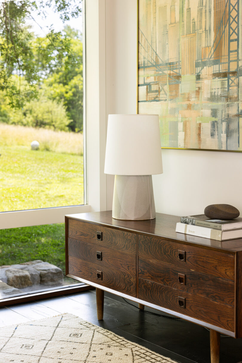 A mid-century modern wooden console with a white table lamp, books, and decor stands by a window overlooking a grassy field. A painting hangs above the console