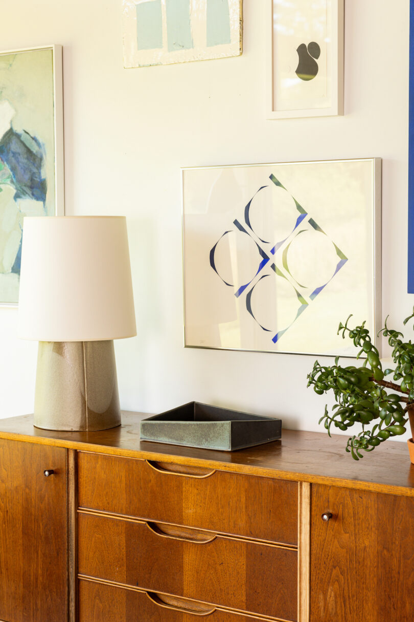 A wooden sideboard with a gray lamp, a plant, and a tray. Abstract artwork and paintings hang on the wall above it