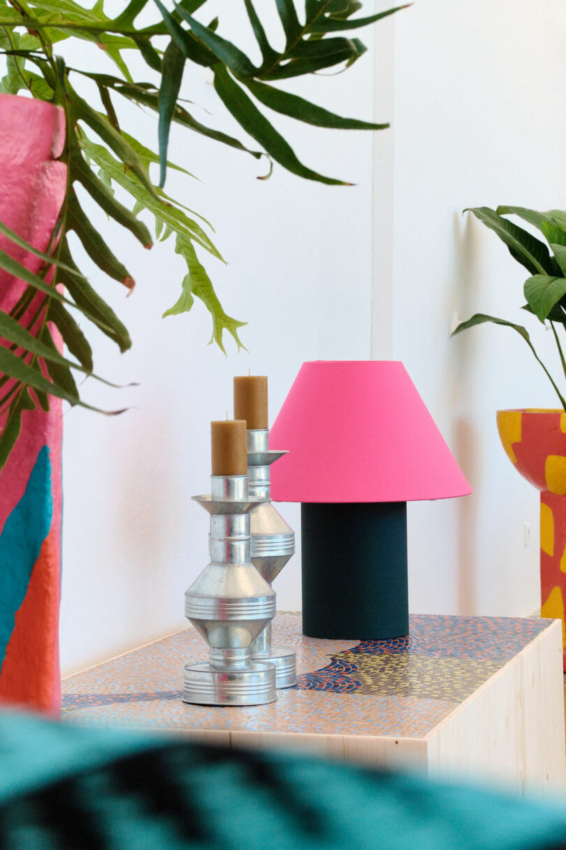 A pink lamp and silver candleholders on a colorful patterned table, surrounded by green plants