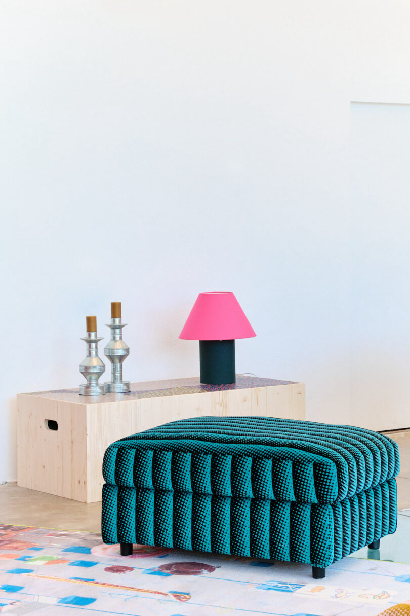 A teal upholstered ottoman is in front of a light wood table with two silver candlesticks and a pink lampshade on a black base. A colorful rug is partially visible on the floor