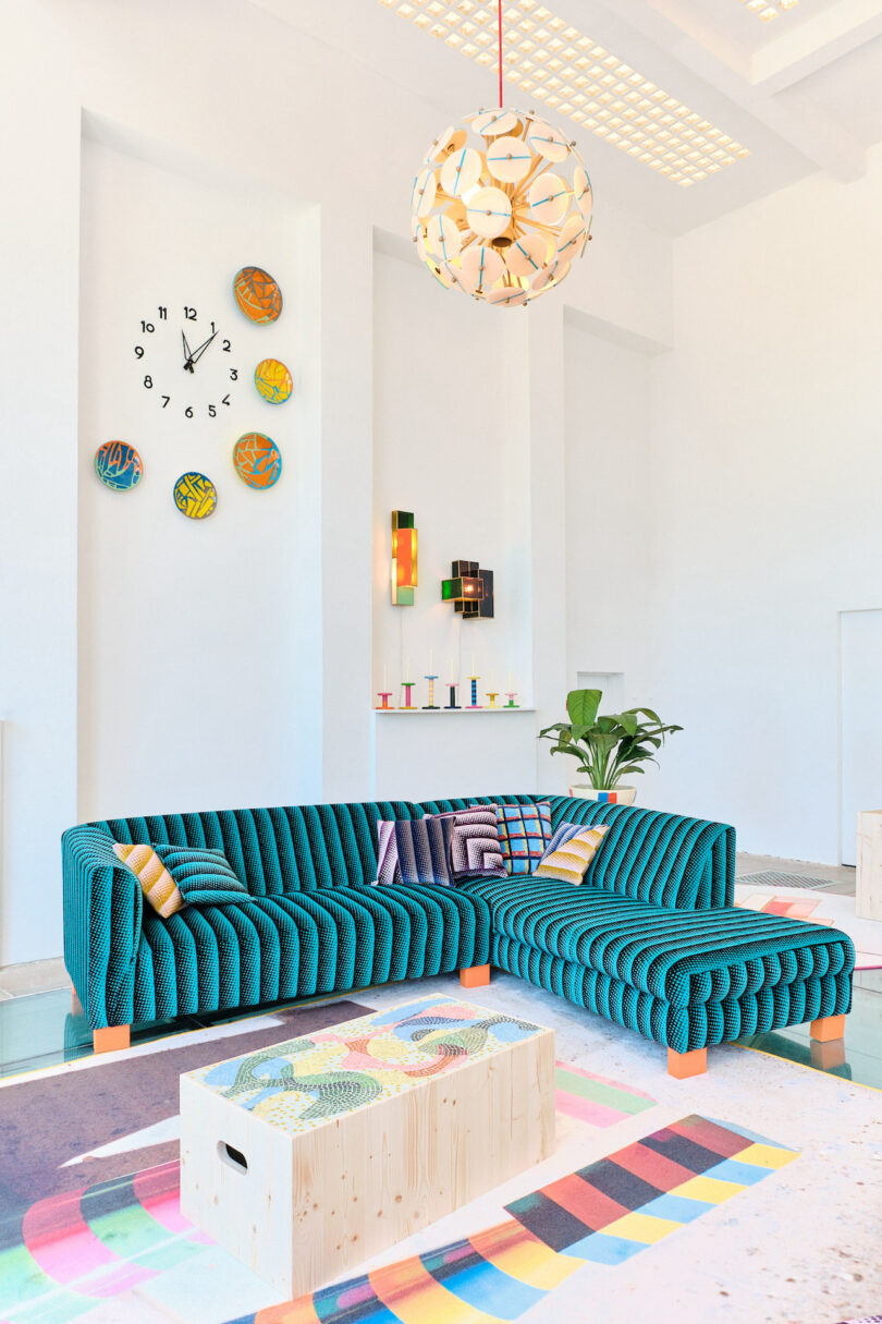 A modern living room with a teal sectional sofa, colorful wall clock, geometric light fixture, and a wooden coffee table with patterned decor. A potted plant is in the corner