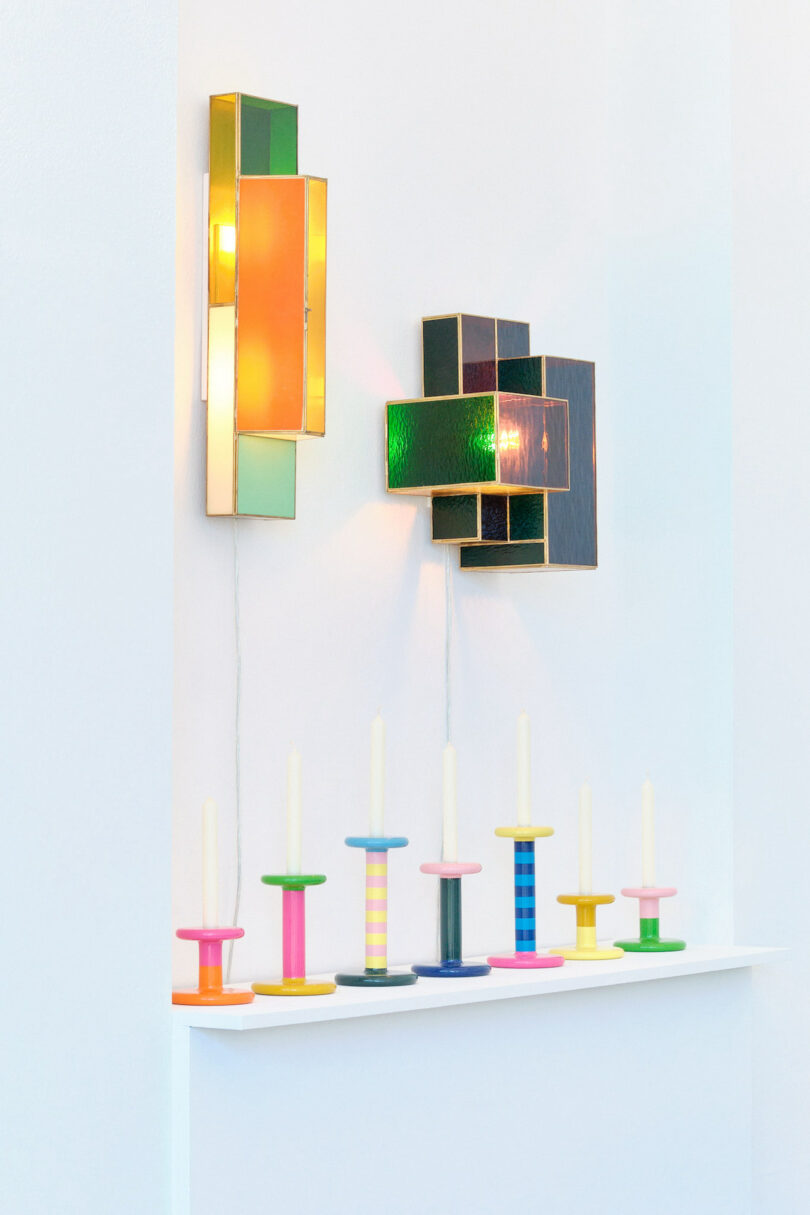 Colorful geometric wall lights and vibrant candlestick holders on a white shelf against a white wall