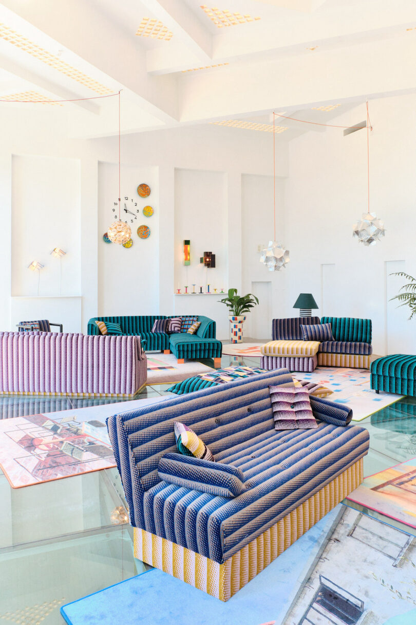 Bright, modern living room with colorful striped sofas, geometric rugs, and decorative wall art. Large windows allow natural light, and contemporary lamps hang from the ceiling