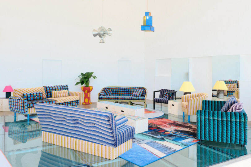 A colorful, modern living room with patterned sofas, a glass coffee table, mismatched lamps, and a plant. Geometric decor and a patterned rug complete the look