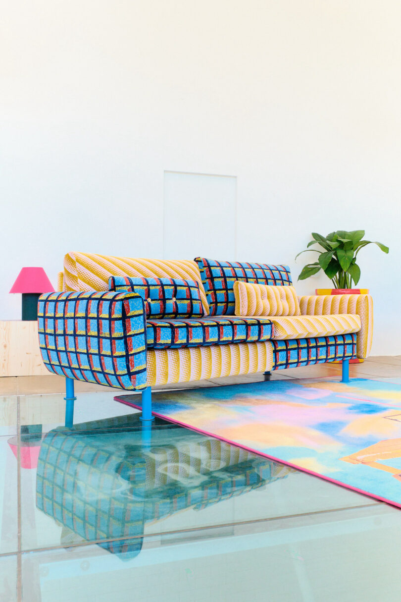 A colorful patterned sofa on a vibrant rug with a pink lamp and green plant in the background