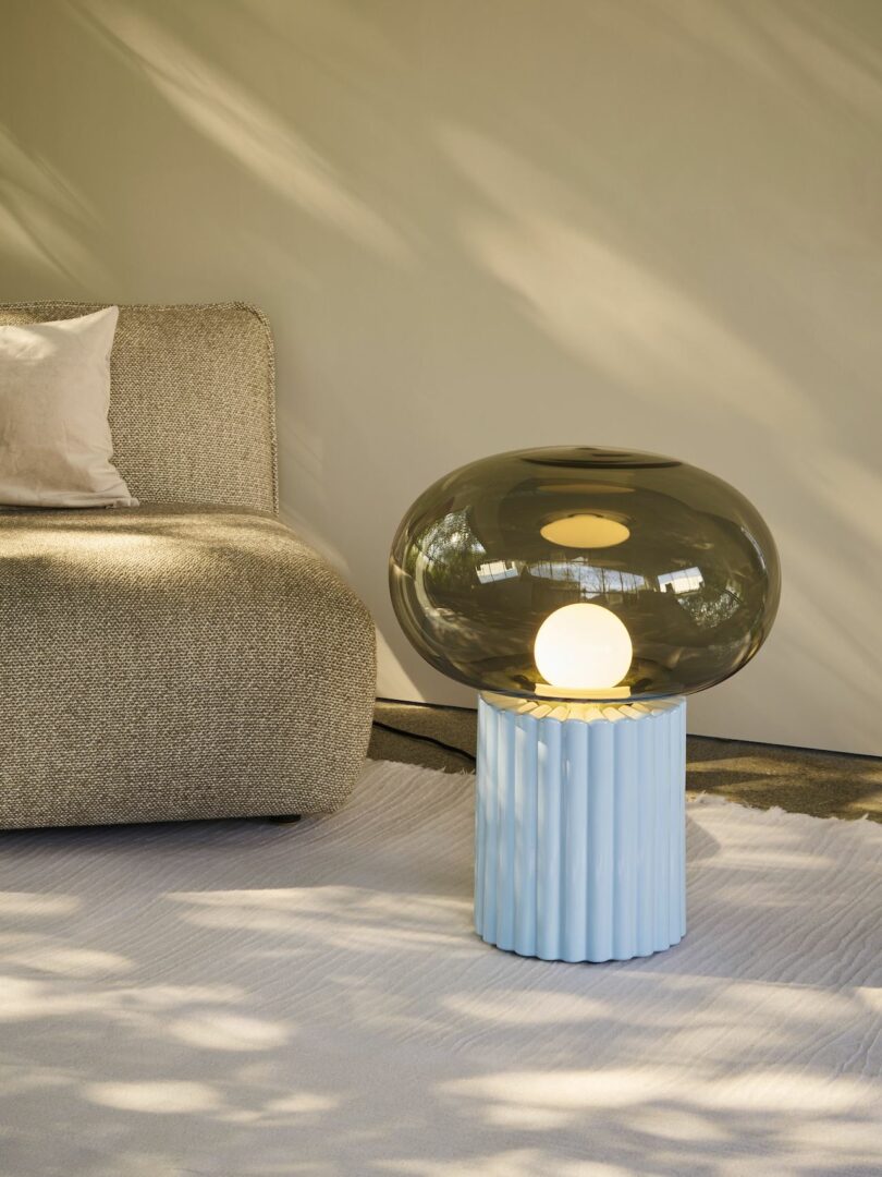 A modern lamp with a round base and globe-shaped shade stands on the floor next to a textured beige sofa. Natural light casts tree shadows on the wall and carpet