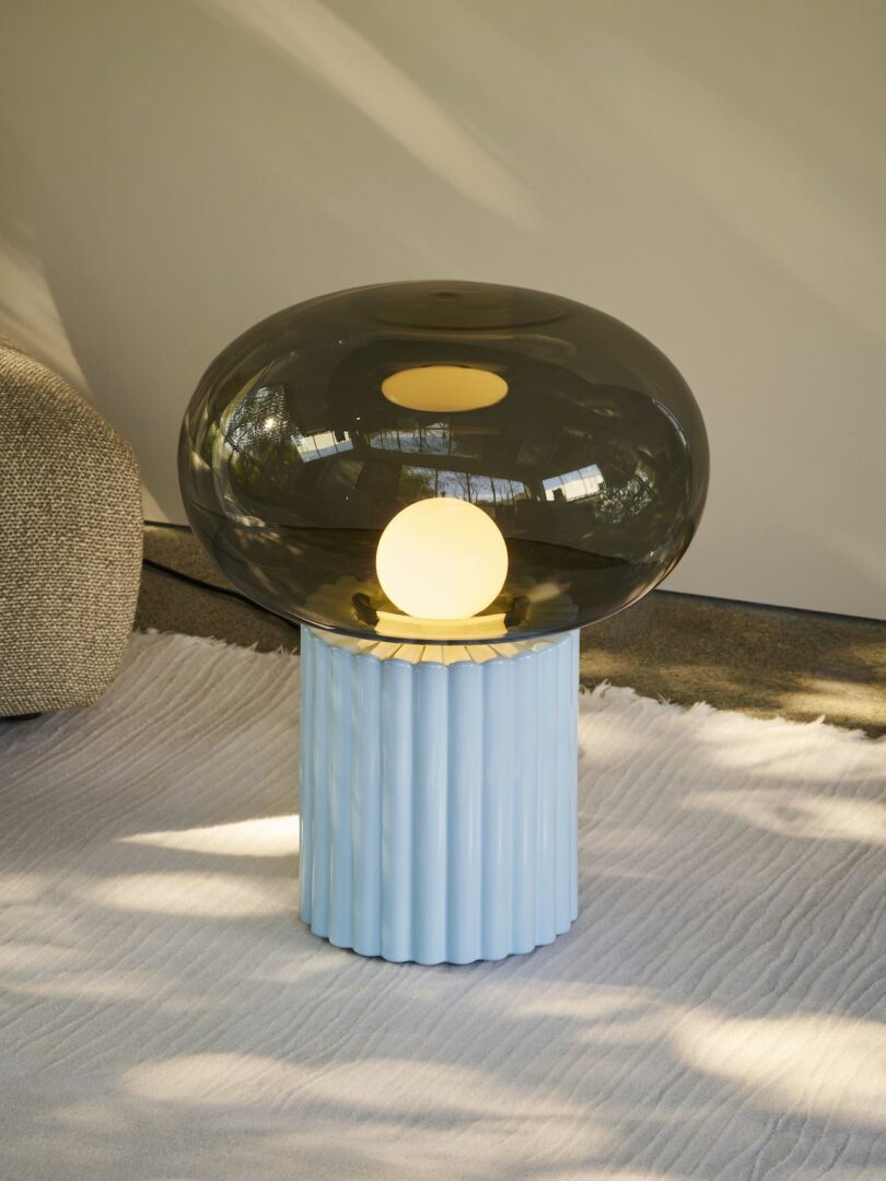 A modern lamp with a translucent dark dome and glowing orb sits on a ribbed blue base, placed on a textured white rug near a beige sofa