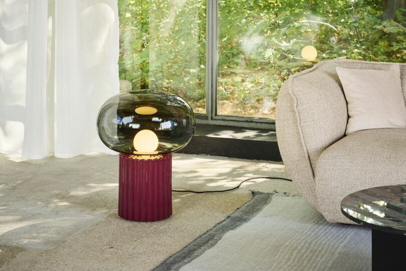 A modern lamp with a round glass shade on a red base sits next to a beige sofa in a bright room with large windows