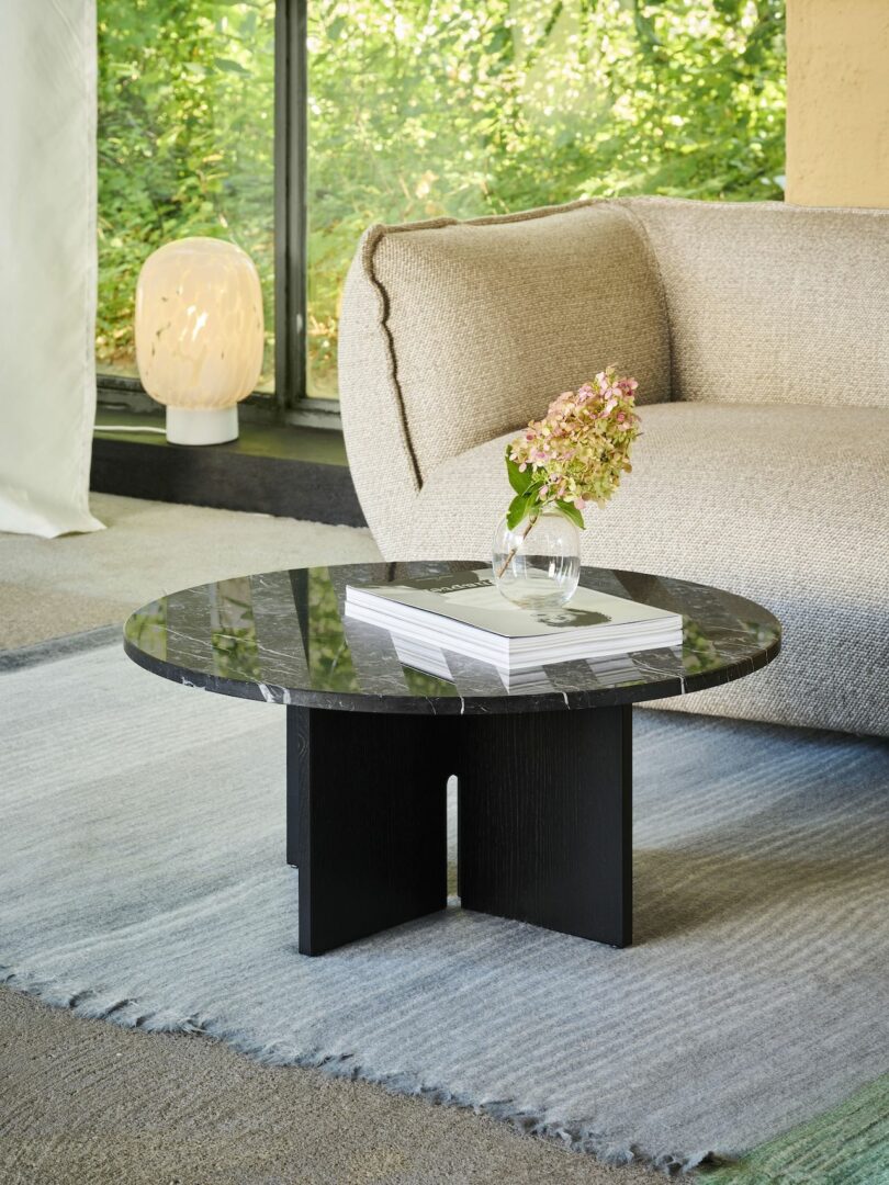 A cozy living room with a beige sofa, round black marble coffee table holding a vase with flowers and books, large window with greenery visible outside, and a lamp in the corner