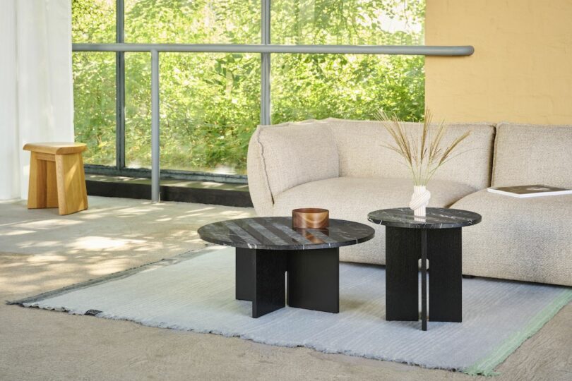 Modern living room with a beige sofa, two black round coffee tables, a wooden stool, and a vase with dried grass. Large windows provide natural light with greenery outside