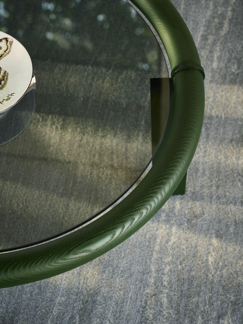 Close-up of a round glass table with a textured dark green frame. Part of a decorative plate is visible on the table. The surface beneath the table has a textured appearance