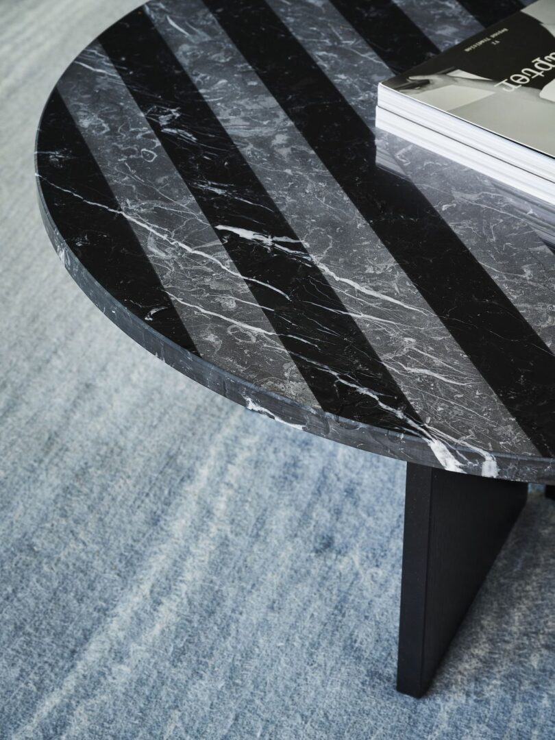 Round marble table with black and gray stripes, set on a light blue carpet, with a stack of magazines on top