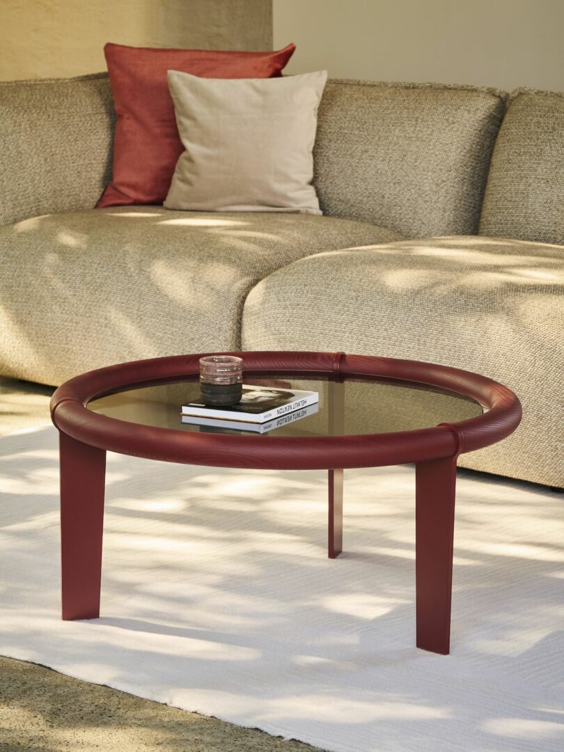 A modern living room features a curved beige sofa with two pillows and a round glass-top coffee table with a red frame on a white rug. A candle and book are on the table