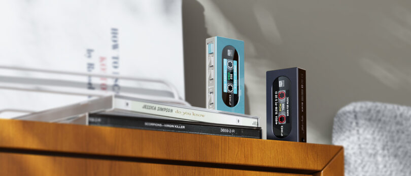 Two compact audio devices resembling cassette tapes rest on a wooden surface, next to stacked CDs.