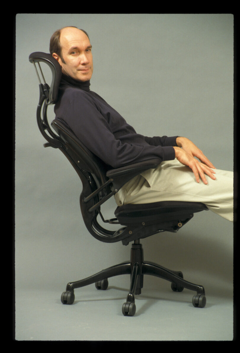 A person reclines in a modern office chair on a plain background, wearing a dark shirt and light-colored pants